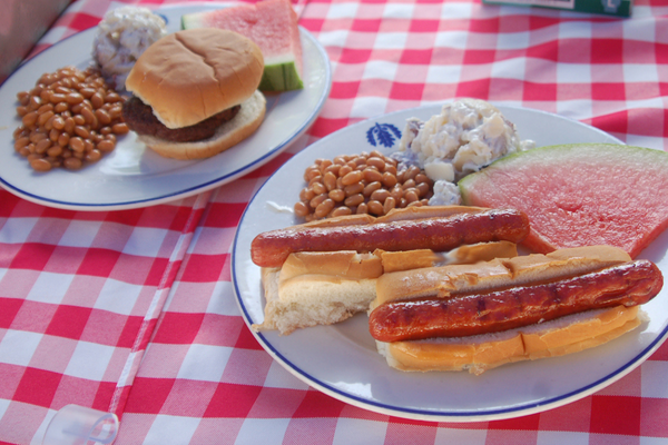 plate with burger, beans, potato salad, & watermelon and 2nd plate with hot dogs, beans, watermelon, and potato salad